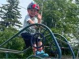 A young boy wearing a helmet on the top of a playground, waving and smiling.