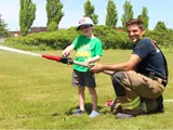 A firefighter is crouching down and helping a small child hold a fire hose, which is spraying a big jet of water on a lawn