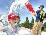 A young boy is smiling, covered in foam, with a firefighter standing behind with a hose spraying foam
