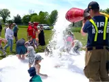 A group of children and adults play in a pile of foam, with a firefighter holding the fire hose and spraying foam just beside them